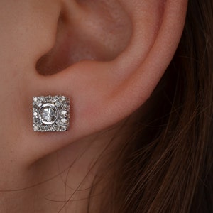 May include: A square-shaped stud earring with a central oval-cut gemstone, surrounded by smaller, round gemstones. The earring is set in a silver or white metal, and is shown on an ear.