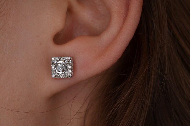 May include: A square-shaped stud earring with a central oval-cut gemstone, surrounded by smaller, round gemstones. The earring is set in a silver or white metal, and is shown on an ear.