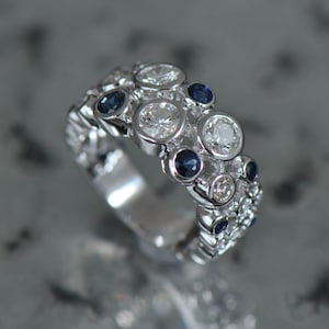 May include: A silver ring featuring a cluster of round, bezel-set diamonds and sapphires. The ring has a textured band and a mix of large and small gemstones, creating a sparkling, eye-catching design. The ring is a piece of jewelry.