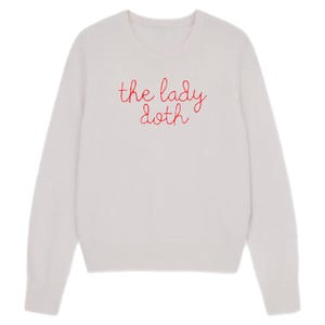 May include: A light-colored sweater with long sleeves. The text "the lady doth" is embroidered in red across the chest. The sweater has a crew neck and a simple, classic design.