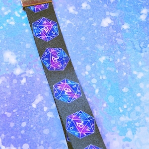 May include: A black fabric wristlet keychain featuring a repeating pattern of twenty-sided dice in shades of purple, blue, and pink. The strap is attached to a silver-toned metal clasp and hardware, designed for holding keys or small accessories.