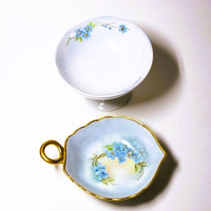 May include: Two small porcelain bowls with blue forget-me-not flower designs. One bowl is on a pedestal and the other is a leaf-shaped bowl with a gold handle.