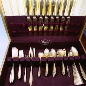 May include: A vintage silver-toned flatware set in a burgundy velvet-lined case. The set includes knives, forks, and spoons, neatly arranged in individual slots. The case is open, revealing the silverware against a gold and cream interior.
