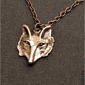 May include: A silver-coloured fox pendant necklace featuring a detailed fox head design. The pendant hangs from a delicate chain. The necklace is displayed on a textured, dark grey background.