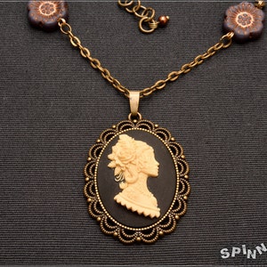 以下が含まれることがあります： ヴィンテージスタイルのカメオネックレス。クリーム色の横顔が黒い背景に映え、楕円形の装飾的なブロンズ色のフレームに収められています。ネックレスは、花型のビーズとマッチするペンダントが付いたブロンズチェーンです。