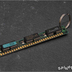 Könnte beinhalten: Ein grüner Schlüsselanhänger mit Leiterplatte, goldenen Anschlüssen und drei schwarzen Chips. Die Chips tragen Aufschriften wie "SIEMENS" und "FRANCE". Ein silberner Schlüsselring ist angebracht. Der Text "SX 0014 7142" ist sichtbar.