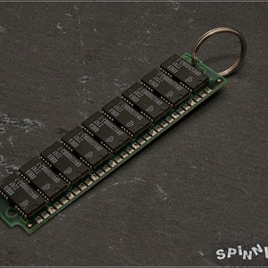 May include: A computer memory module keychain with a silver key ring. The green circuit board holds several black rectangular chips. The keychain is set against a dark gray textured surface.