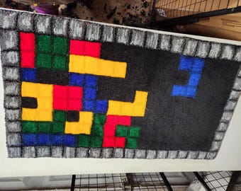 May include: A colorful rug with a Tetris-like design. The rug is made of a soft, plush material and features a variety of colors, including red, yellow, blue, green, and black. The rug is surrounded by a gray border.