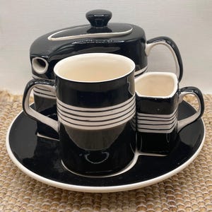 Puede incluir: Juego de té en blanco y negro con tetera, dos tazas y una pequeña cremaera en una bandeja a juego. El juego presenta rayas blancas horizontales sobre fondo negro.
