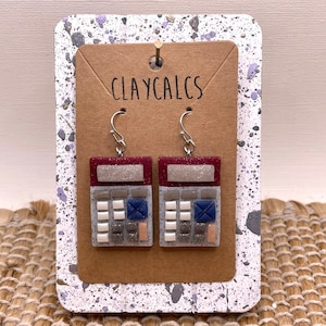 May include: A pair of red and white calculator earrings with silver hooks. The earrings are made of polymer clay and have a glittery finish. The text "CLAYCALCS" is printed on the card behind the earrings.