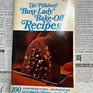 May include: A vintage Pillsbury Busy Lady Bake-Off Recipes cookbook featuring a slice of chocolate cake on the cover. The title is printed in blue text, and the cover includes the text 50 cents and 100 prizewinning recipes shortcuted and up-to-dated.