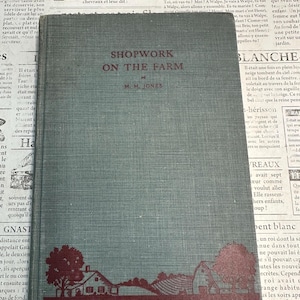 May include: A gray hardcover book titled Shopwork on the Farm by M. M. Jones. The cover features a dark red illustration of a farmstead with trees and a house at the bottom, and the text McGraw-Hill Rural Activities Series in a red banner.