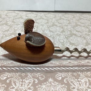 May include: A novelty corkscrew shaped like a mouse. The wooden body is brown with black eyes and brown and gray ears. The corkscrew is made of silver-colored metal.
