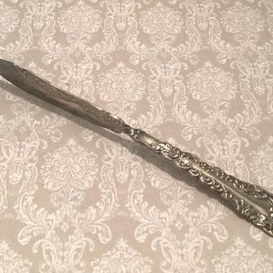 May include: Silver butter knife with a decorative floral pattern on the handle and a pointed blade. The knife is set against a patterned background. This vintage utensil is ideal for serving butter or other spreads.