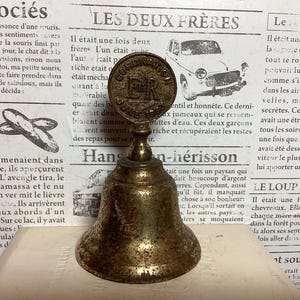 May include: Vintage brass bell with a detailed design. The bell features a round top with the text "CORONATION JUNE 1953 ER" and a bell-shaped bottom. The bell is set against a backdrop of old newspaper.