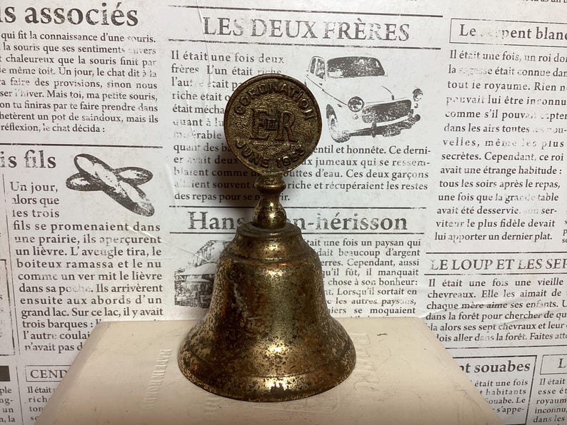May include: Vintage brass bell with a detailed design. The bell features a round top with the text "CORONATION JUNE 1953 ER" and a bell-shaped bottom. The bell is set against a backdrop of old newspaper.