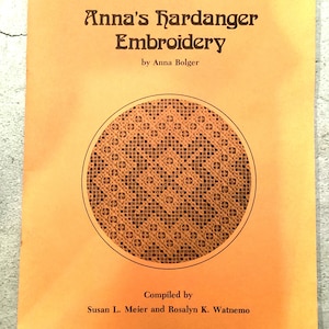 May include: An orange book cover with the title "Anna's Hardanger Embroidery" and a circular embroidery design. The author is Anna Bolger, and the book is compiled by Susan L. Meier and Rosalyn K. Watnemo.