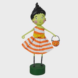 May include: A Halloween-themed figurine of a girl with green skin, black hair, and an orange and white striped dress. She holds a pumpkin-shaped basket and stands on a green base. The figurine has a surprised expression and is wearing a yellow bow.