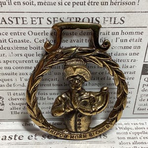 Pode incluir: Um emblema decorativo de metal dourado com uma figura com um chapéu de chef segurando uma trompa, rodeada por uma coroa de louros. O texto "RIPON HORN BLOWER" está inscrito na parte inferior. O emblema está sobre um fundo de papel envelhecido.