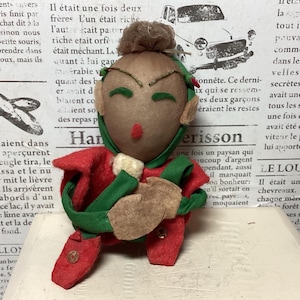 Könnte beinhalten: Handgefertigte Stoffpuppe in roter und grüner Kleidung. Das Gesicht ist braun, mit grünen Augen, einer roten Nase und einem kleinen braunen Dutt. Die Puppe sitzt mit verschränkten Armen.