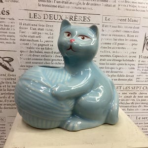 Puede incluir: Figura de gato de cerámica azul claro, sosteniendo una bola de lana. El gato tiene ojos rojos y nariz rosa. La figura es un objeto decorativo, posiblemente una maceta o un adorno para el hogar.