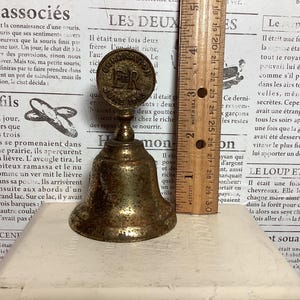 May include: Antique brass bell with a round top featuring the text "CORONATION JUNE 1937 ER". The bell is approximately 3 inches tall and sits on a white surface. The bell is a vintage collectible.