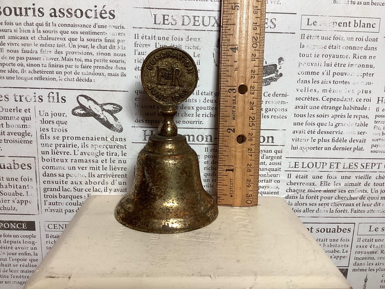 May include: Antique brass bell with a round top featuring the text "CORONATION JUNE 1937 ER". The bell is approximately 3 inches tall and sits on a white surface. The bell is a vintage collectible.