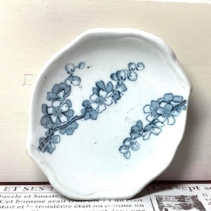 May include: A small, white, ceramic dish with an irregular shape. The dish is decorated with a blue floral pattern. The flowers are arranged on branches, creating a delicate and artistic design. The dish is likely used for serving small portions.