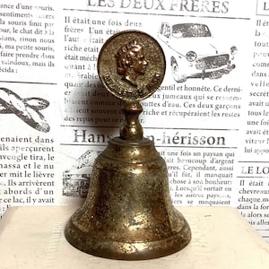 May include: Antique brass bell with a detailed coin-like top featuring a profile. The bell has a flared base and a tarnished finish, suggesting age. The background includes vintage French text.