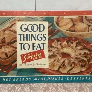 May include: A retro cookbook cover with the title "Good Things to Eat" in blue. The cover displays images of baked goods, including pies, biscuits, and a cinnamon roll. The text "My Surprise Recipe" is also visible. The bottom of the cover reads "Hot Breads Meat Dishes Desserts".