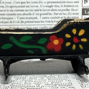 May include: A small, black wooden sleigh with a curved back and rocking base. The sleigh is decorated with red, yellow, and green floral designs and gold trim. A charming decorative piece.