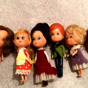 Könnte beinhalten: Eine Sammlung von sechs Vintage-Puppen mit verschiedenen Frisuren und Outfits. Die Puppen haben bemalte Gesichter und sind in verschiedenen Farben gekleidet, darunter Rot, Weiß, Grün und Lila. Einige Puppen tragen Kleider, andere sind unbekleidet.