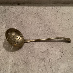 May include: A vintage, brass-coloured slotted spoon with a decorative floral pattern in the bowl. The handle is curved and slender. The spoon is designed for straining liquids, likely used in cooking.