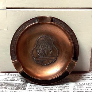 May include: Vintage copper ashtray with a Native American chief profile in the center. The ashtray has a circular shape with a decorative border and three notches for cigarettes. The text "Fortunes of War" is visible.