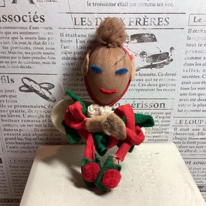 Könnte beinhalten: Eine handgefertigte Vintage-Puppe mit einem hellbraunen Gesicht, blauen Augen und einem roten Mund. Die Puppe hat einen braunen Dutt, rote und grüne Kleidung und ist vor dem Hintergrund einer alten Zeitung drapiert.