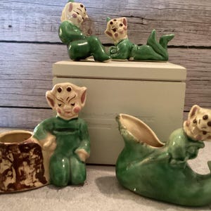 May include: A collection of vintage ceramic elf figurines in various poses. The elves are primarily green with cream-colored faces and accents. One elf is seated, another reclines, and two are planters shaped like a boot and a tree stump.