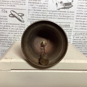 May include: A vintage brass bell with a clapper, resting on a white surface. The bell has a rounded shape and a tarnished finish, suggesting age. The background features newspaper print.