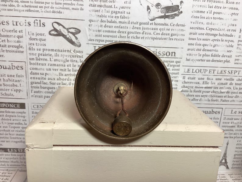 May include: A vintage brass bell with a clapper, resting on a white surface. The bell has a rounded shape and a tarnished finish, suggesting age. The background features newspaper print.