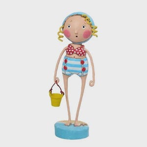 May include: A whimsical figurine of a beachgoer. The figure wears a blue swim cap, a red polka-dot bikini top, and blue and white striped shorts with red buttons. She holds a yellow bucket with a red handle. The figurine stands on a blue base.