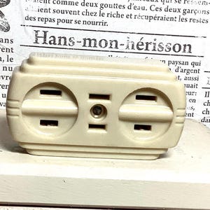 Puede incluir: Un enchufe eléctrico vintage de color blanquecino con dos enchufes estándar y una conexión de tierra central. El enchufe rectangular tiene bordes redondeados. Probablemente está hecho de baquelita o un material similar. Un diseño clásico.