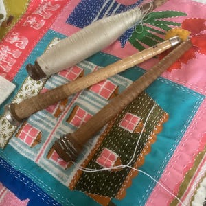 May include: Three wooden spinning bobbins, one wrapped in white thread and two empty. The bobbins feature cylindrical shafts with metal ends and varying wood tones, ranging from light tan to a darker brown, arranged diagonally on a colourful fabric.