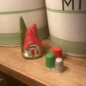 May include: A small red and white ceramic house with a green tree, three thimbles, one red, one green, and one silver.