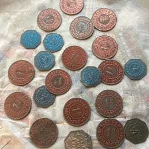 May include: A selection of vintage metal tokens in various shapes and sizes. The tokens are mainly red and blue, with text reading "HINCKLEY CO-OP LIMITED" and denominations like "1P" and "1/2D". The tokens are arranged on a light-coloured surface.
