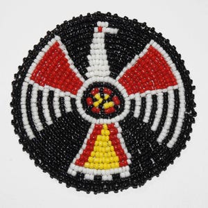 May include: Round beaded emblem featuring a stylized bird design. The emblem is primarily black, with red, white, and yellow accents. The bird's wings and body are detailed with small, colorful beads, creating a textured appearance.