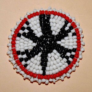 May include: Circular beaded design featuring a central black cross shape surrounded by white beads. A ring of red beads borders the design. The craftwork is likely a pin or patch, showcasing a Native American or similar cultural motif.