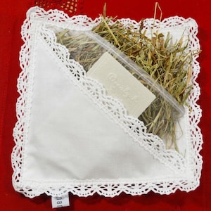 Puede incluir: Una bolsa de tela blanca con un borde de encaje, que contiene una pequeña barra de jabón blanca y heno seco en una bolsa de plástico transparente. La bolsa tiene una pequeña etiqueta con la palabra "nan" en ella.