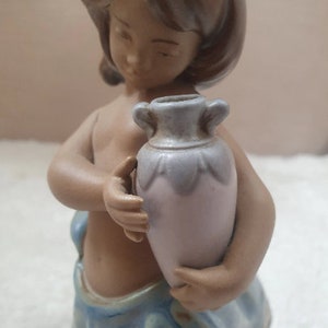 May include: A ceramic figurine of a girl holding a light blue and white pitcher. The girl is wearing a blue and white dress and has brown hair.