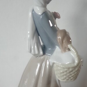 Pode incluir: Uma figura de porcelana de uma mulher usando um len&ccedil;o branco na cabe&ccedil;a e um vestido azul, segurando uma cesta com uma al&ccedil;a tecida.