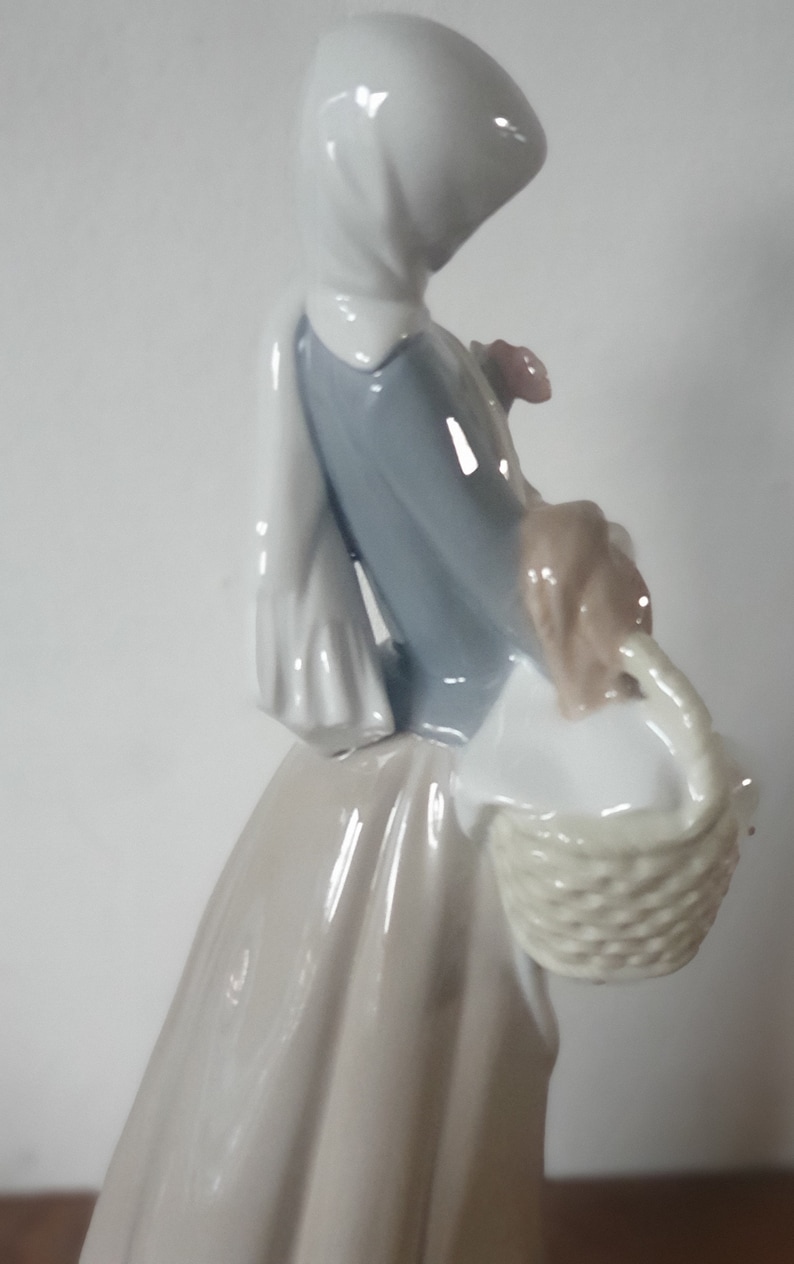 Pode incluir: Uma figura de porcelana de uma mulher usando um len&ccedil;o branco na cabe&ccedil;a e um vestido azul, segurando uma cesta com uma al&ccedil;a tecida.