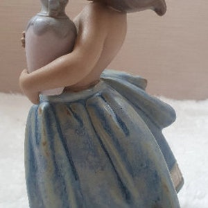 May include: A porcelain figurine of a girl wearing a blue dress and holding a pink and white pitcher.